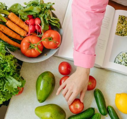 Top view of fresh vegetables and hands arranging them with a recipe book, promoting healthy eating.