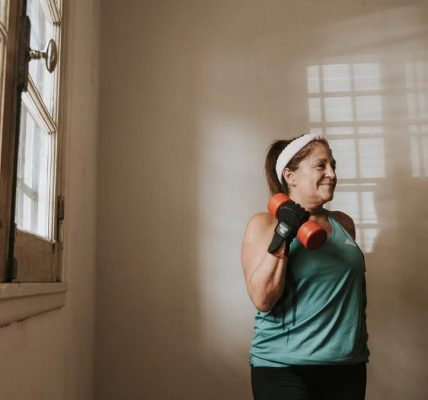 Senior woman in a brightly lit room exercises with dumbbells, promoting fitness at home.