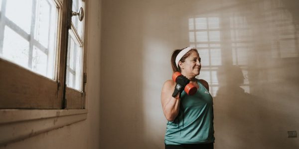 Senior woman in a brightly lit room exercises with dumbbells, promoting fitness at home.